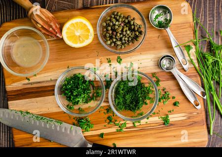 Erbe, capperi e limone tritati su un tagliere di legno: Aneto fresco, capperi, prezzemolo e succo di limone su un tagliere di legno Foto Stock