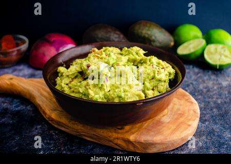 Guacamole fresche in una ciotola di legno con ingredienti sullo sfondo: Guacamole fatte in casa con avocado, lime, cipolla, coriandolo e peperoni Foto Stock