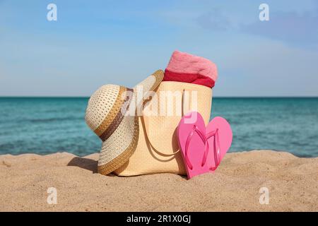 Borsa estiva con pantofole, telo da spiaggia e cappello di paglia sulla sabbia vicino al mare Foto Stock