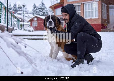 ritratto uomo latino con baffi e giacca con cappuccio, abbracciando un cane san bernardo insieme in profilo nella neve d'inverno in un complesso di cabina. Orizzontale y Foto Stock
