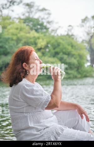 Donna ispanica matura seduta in un parco bere succo verde da una bottiglia di vetro. Concetti: Nutrimento sano e stile di vita. Foto Stock