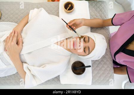 Giovane donna asiatica con nutriente viso vestito in accappatoio, ricevendo cura del viso da estetista al salone termale, Top View. Maschera peeling viso, bellezza spa tr Foto Stock