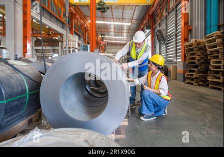 Il Senior Engineer Manager forma i nuovi dipendenti all'interno della fabbrica di lamiere. Tutti indossano giubbotto di sicurezza e elmetto. La pila di rulli in lamiera di acciaio si trova in Foto Stock