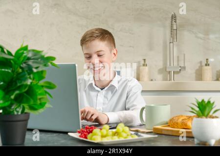 giovane ragazzo, adolescente gioca sul notebook e sul computer portatile durante la colazione. Dipendenza dai gadget. Foto Stock