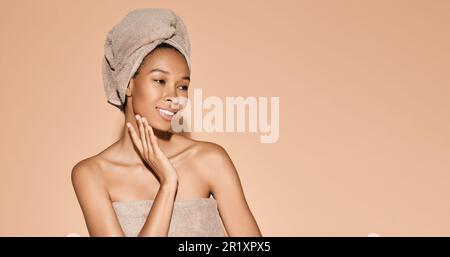 Concetto di cura del corpo. Donna afro-americana felice con pelle fresca idratata con asciugamano sulla testa dopo le procedure di bagno su sfondo beige Foto Stock