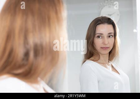 Ritratto di affascinante bella Brunette Caucasian 40 Yo Donna guardando Specchio e sorridente. Salute idratata cura della pelle, invecchiamento bellezza, Skincare Foto Stock