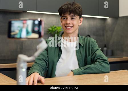 Giovane uomo sorridente blogger influencer sparare se stesso su uno smartphone su uno stabilizzatore in cucina Foto Stock