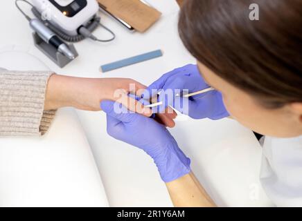 L'estetista del chiodo ritagliato rimuove la cuticola del cliente femminile con il bastone arancione, vista dall'alto. Lima, attrezzi sterilizzati per manicure in confezione artigianale su tavolo bianco Foto Stock