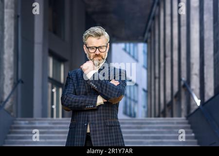 Ritratto di serio pensando uomo d'affari maturo, uomo grigio più anziano con capelli in tuta d'affari fuori ufficio edificio pensando e guardando al lato, investitore esperto banchiere con barba. Foto Stock