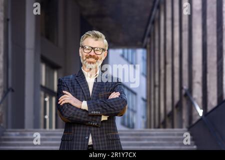 Ritratto di uomo d'affari maturo grigio-haired, capo senior sorridente e guardando la macchina fotografica con le braccia incrociate, uomo grigio-haired al di fuori della costruzione dell'ufficio, investitore riuscito e proprietario in vestito di affari Foto Stock
