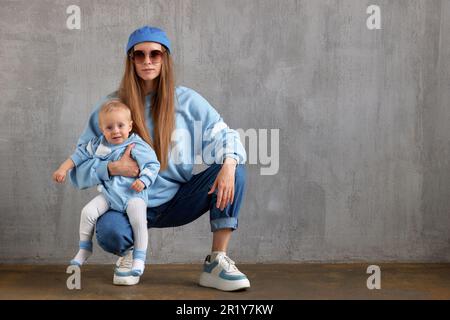 Giovane mamma affascinante abbraccia il suo prezioso bambino. Il bambino felice siede sul grembo della madre. Entrambi sono vestiti con abiti bianchi e blu simili. La felicità della famiglia An Foto Stock