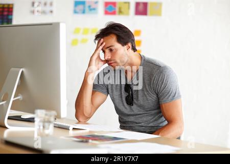 Sofferenza da scrittori blocco. Un bell'uomo seduto alla scrivania e guardando lo schermo di un computer con la testa appoggiata sulla mano. Foto Stock