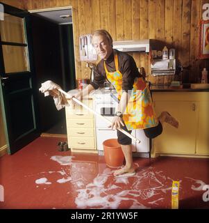 Giorno di pulizia nel 1970s. Un interno di una cucina dove si vede un uomo che pulisce il pavimento. Per velocizzare il lavoro, ha montato delle spazzole sui piedi. È attore Stig Grybe 1928-2017. 4 ottobre 1972. Kristoffersson Foto Stock