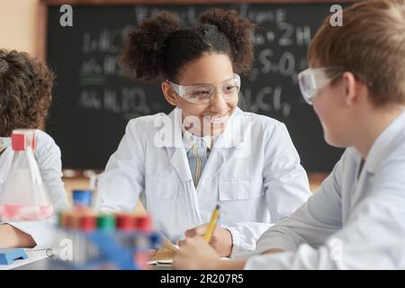 Ritratto di una studentessa nera sorridente che indossa un camice da laboratorio in classe scientifica e parla con un amico. Foto Stock