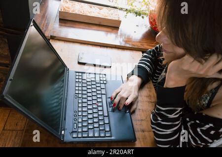 Profilo primo piano di una giovane donna transgender Latina di etnia argentina, utilizzando il suo laptop in un ristorante, navigando in internet, controllando le e-mail Foto Stock