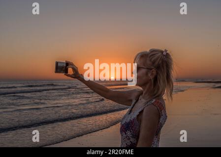 Donna matura sorridente mentre si scattano selfie con un telefono cellulare sulla spiaggia al tramonto. Foto Stock