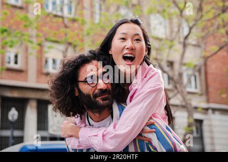 Giovane coppia multirazziale felice che si diverte abbracciando e abbracciando toguether. Il marito caucasico sorrideva e regalava un giro in piggyback alla sua bella moglie asiatica in una data romantica o in luna di miele. Foto di alta qualità Foto Stock