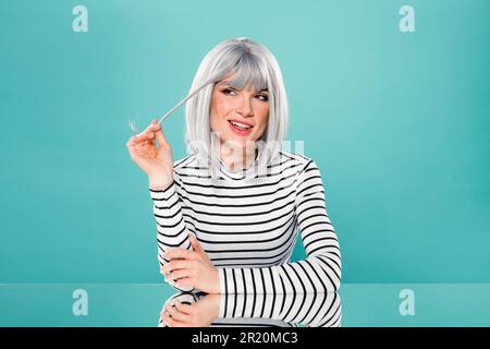 Ritratto di positiva giovane donna in abito alla moda sorridente e guardando lontano mentre si siede su sfondo turchese Foto Stock