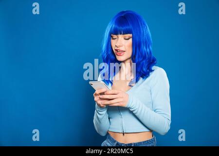 Positivo giovane donna con capelli tinti blu in abiti casual navigazione cellulare su sfondo blu Foto Stock