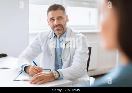 Assistenza incentrata sul paziente. Allegro medico maschile consulenza signora paziente, compilando il modulo di consultazione in clinica Foto Stock