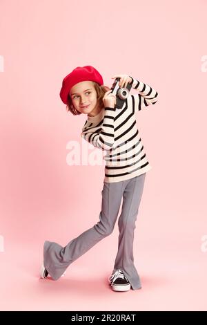 Ritratto di curiosa bambina, cild in maglione a righe e berretto rosso posa con vintage foto camra agaisnt sfondo rosa studio Foto Stock