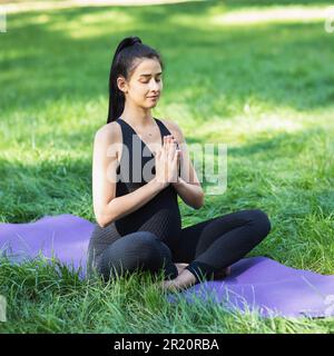 Giovane donna incinta che conduce uno stile di vita sano, meditando, seduta in una posizione di loto con namaste, facendo yoga su un tappetino nel parco in una spo nera Foto Stock