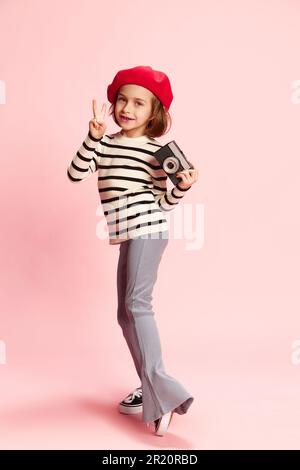 Ritratto di piccola ragazza positiva, cild in maglione a righe e berretto rosso posa con foto vintage camra agaisnt sfondo rosa studio Foto Stock