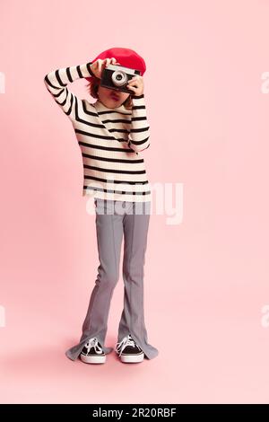 Ritratto di bambina, cild in maglione a righe e berretto rosso posa con foto vintage camra agaisnt sfondo rosa studio. Turistico Foto Stock