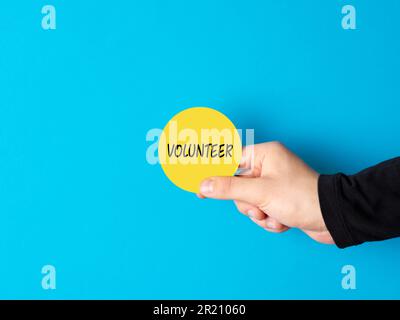 Concetto di volontariato. Aiuto, assistenza, beneficenza e comunità di sostegno. Mano maschile che tiene un segno di cerchio giallo con la parola volontario contro bac blu Foto Stock