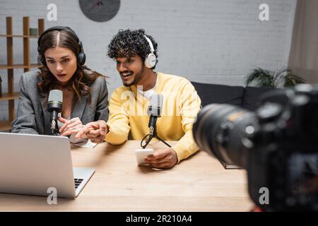 allegro uomo indiano in cuffie e jumper giallo che tiene lo smartphone e indica il portatile vicino stupito collega brunette e fotocamera digitale su blu Foto Stock