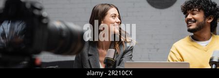 uomo indiano barbuto in jumper giallo e affascinante bruna donna in blazer grigio sorridente l'un l'altro mentre registra podcast di fronte allo studio microp Foto Stock