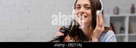 Bruna podcaster sorridente e toccante cuffie wireless che tiene il microfono e guardare la fotocamera durante lo streaming in podcast in studio, banner, stock Foto Stock