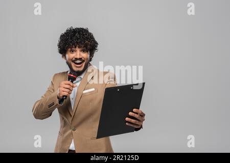 Allegro e giovane ospite indiano di evento con capelli ricci in piedi in abiti formali parlando al microfono e tenendo appunti durante la festa isolato su g Foto Stock