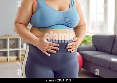 Donna sovrappeso, obesa in reggiseno sportivo e leggings tenendo le mani sul ventre grasso Foto Stock