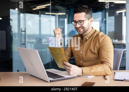 Felice uomo d'affari giovane apre una busta con una lettera al tavolo in ufficio. Ricevuta la buona notizia, gioisce, mostra un gesto di vittoria con il suo Foto Stock