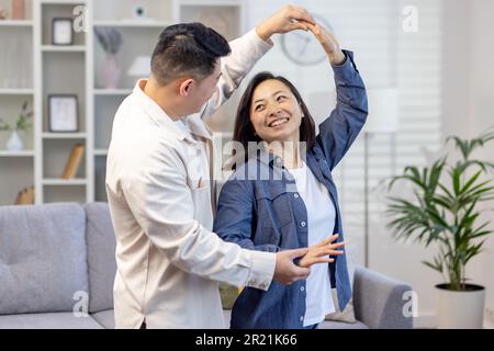 Felice coppia asiatica, giovane famiglia uomo e donna celebrare il trasferimento in una nuova casa, l'acquisto di un appartamento. Ballano, abbracciano, ridono. Foto Stock