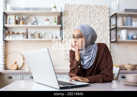 Annoiava una giovane donna musulmana che indossava un hijab seduto in cucina a casa, appoggiava la testa sulle mani, e guardava triste e moody il laptop. Foto Stock