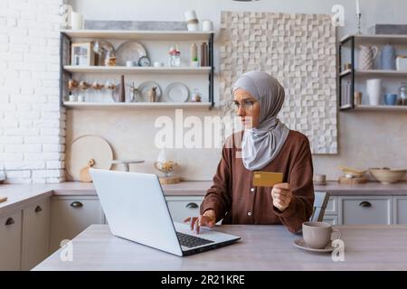 Una giovane donna musulmana che indossa un hijab è seduta in cucina utilizzando un computer portatile e tenendo una carta di credito. Effettua acquisti online, paga fatture, trasferisce fondi, controlla il budget. Foto Stock