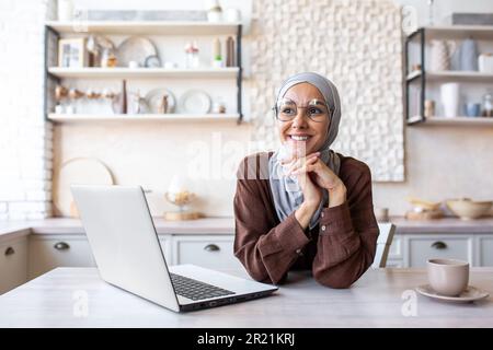 Ritratto di una giovane donna in un hijab seduta sognante e pensieroso in cucina a casa vicino a un computer portatile. Studia online, lavora in remoto, si riposa. Foto Stock