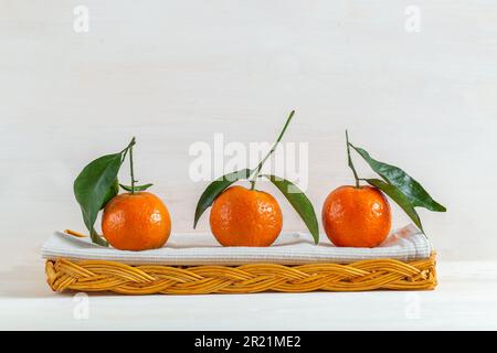 Una vita ferma di tre tangerini freschi, vista frontale, fianco a fianco, in un cesto di vimini con un asciugamano da cucina. I tangerini hanno il loro gambo e le loro foglie Foto Stock