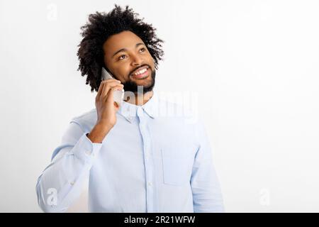 Curly amichevole sorridente felice afroamericano uomo in camicia bianca parlando su cellulare, conducendo piacevole conversazione isolato su sfondo bianco, ritratto studio Foto Stock