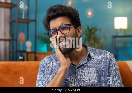 Problemi dentali. Il giovane indiano che tocca la guancia, chiudendo gli occhi con espressione di terribile soffre di dolore ai denti, denti sensibili, cavità. Ragazzo indù a casa appartamento soggiorno sul divano Foto Stock