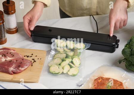 Donna che usa il sigillante per il confezionamento sottovuoto con sacchetto di plastica di zucchine al tavolo bianco, primo piano Foto Stock