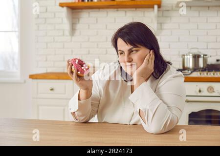 Giovane donna in sovrappeso penosa che esita a mangiare ciambella Foto Stock