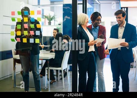 Gruppo di colleghi multirazziali positivi nell'abbigliamento formale che discutono i documenti mentre si trovano contro le pareti di vetro nello spazio di lavoro contemporaneo Foto Stock