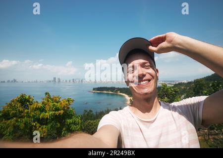 Uomo felice che scatta foto al selfie dal giorno delle vacanze estive. Bel turista che indossa il cappello e sorride alla macchina fotografica contro il paesaggio con la spiaggia. Foto Stock