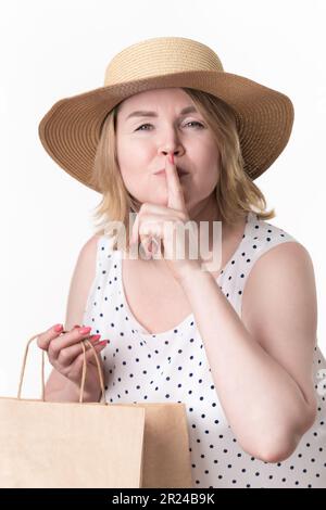 Il cliente femminile serio ha sollevato il suo dito indice alle labbra, fa il segno segreto o chiedendo di mantenere il silenzio e tenendo il sacchetto di shopping del mestiere della carta in altra mano Foto Stock