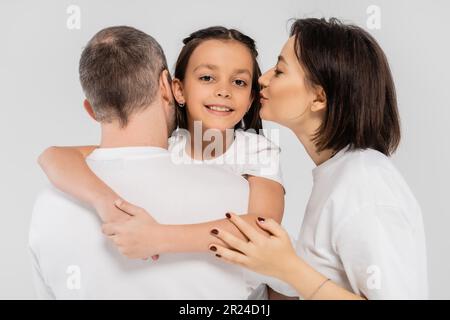 madre con tatuaggio e capelli corti baciando guancia di sorridente figlia preseen vicino al marito mentre sta in piedi in t-shirt bianche insieme su sfondo grigio, Foto Stock