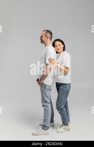 tutta la lunghezza della donna tatuata con i capelli corti della brunetta appoggiati alla parte posteriore del marito sorridente mentre levandosi in piedi insieme nelle t-shirt bianche e nei jeans del denim sopra Foto Stock