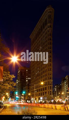 Il Flatiron Building si illumina caldo sotto i semafori sotto un cielo indaco prima dell'alba; il traffico sparso lascia sentieri leggeri nella foto di esposizione al tempo. Foto Stock